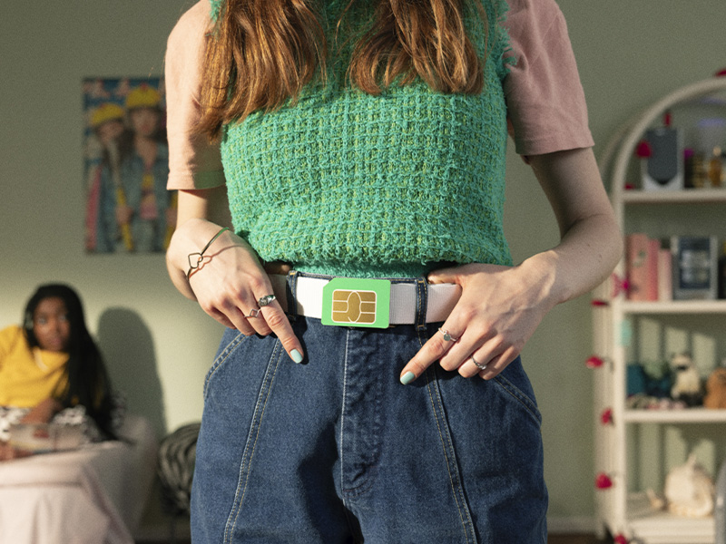 Närbild på en person med grön slipover och jeans som pekar på ett bälte med ett stort SIM-kort – en kreativ symbol för Vimlas mobilabonnemang.